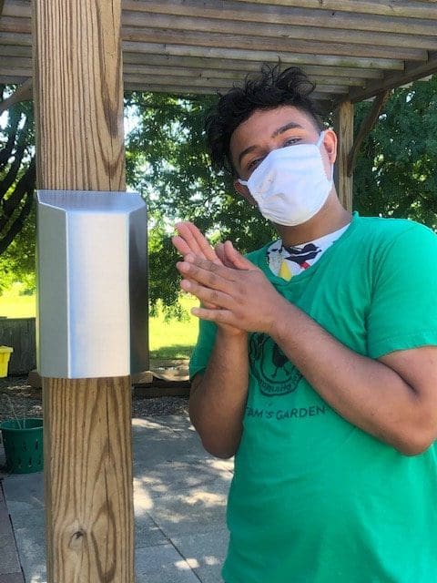 a young person in a green shirt and white mask demonstrates using an automatic hand sanitizer dispenser by rubbing their hands together